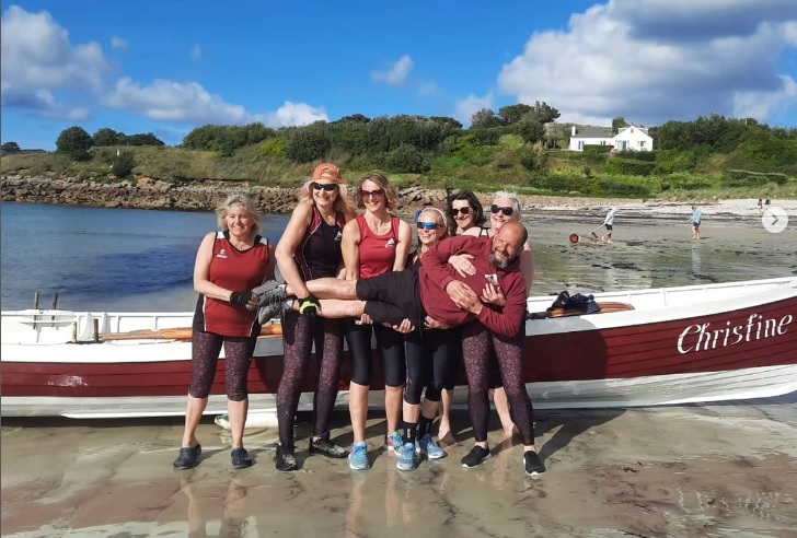 Ladies vets crew in front of the gig on the beach, holding their cox horizontally